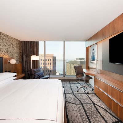 A modern hotel room with a large bed, wall-mounted TV, desk, chair, and window view of buildings and water.