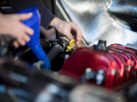 Someone is performing car maintenance, using a blue funnel to add fluid to the engine, with a focus on the engine's red parts.