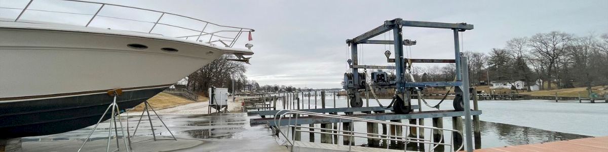 A large white boat is lifted on a dock by a sling, with a metal gangway and a wooden platform over calm water at a marina.