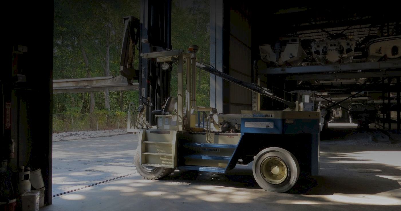 A large blue forklift parked inside a dim industrial warehouse, with sunlit concrete floors and a shadowy doorway behind it.