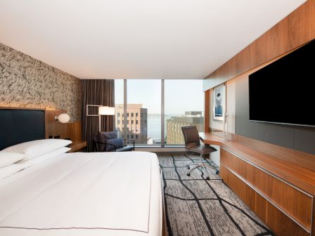 A modern hotel room with a large bed, wall-mounted TV, desk, chair, and a window with a city view.