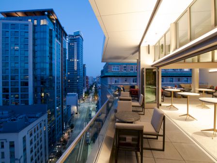 An outdoor terrace at night overlooks a street lined with modern high-rise buildings, featuring chairs, tables, and sleek design.