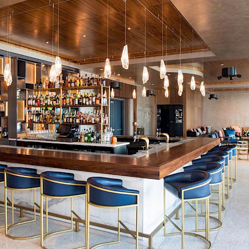 A modern bar with a sleek counter, blue stools, pendant lights, and a stocked back bar is seen. Lounge seating is visible in the background.