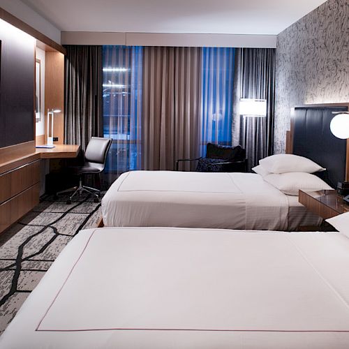 A modern hotel room features two neatly made beds, a wall-mounted TV, a desk with a chair, and stylish lighting and decor, alongside large windows.