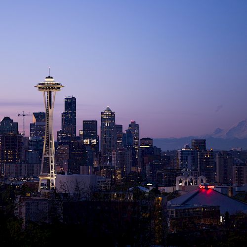 A panoramic view of the Seattle skyline at dusk, prominently featuring the Space Needle with Mount Rainier in the background.